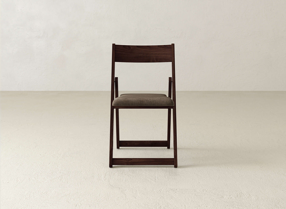 The Emmett Upholstered Folding Chair by James & James, featuring a dark solid maple frame and cushioned seat, sits centered in an empty light room with textured walls and floors.