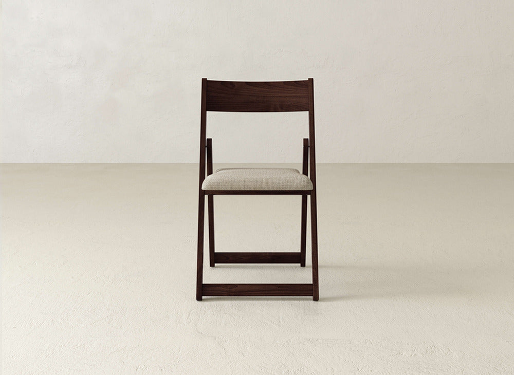 The Emmett Upholstered Folding Chair by James & James features a modern solid maple frame and beige cushioned seat, displayed facing forward against a plain, light wall and floor.