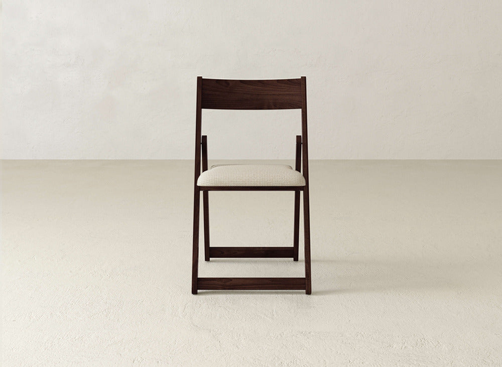 The Emmett Upholstered Folding Chair by James & James features a dark wooden frame, light beige cushioned seat, and foldable design. It is centered on a plain, light floor against a textured off-white wall.