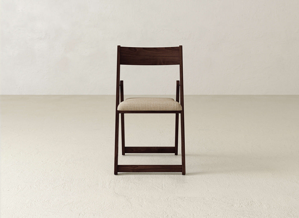 The Emmett Upholstered Folding Chair by James & James, featuring a dark solid maple frame and light beige cushioned seat, is centered on a textured light floor against an off-white wall, facing the camera directly.