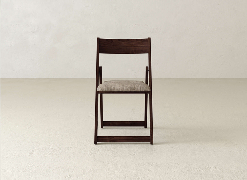 The Emmett Upholstered Folding Chair by James & James, crafted from solid maple hardwood with a padded light beige seat, sits on a light floor against an off-white wall.
