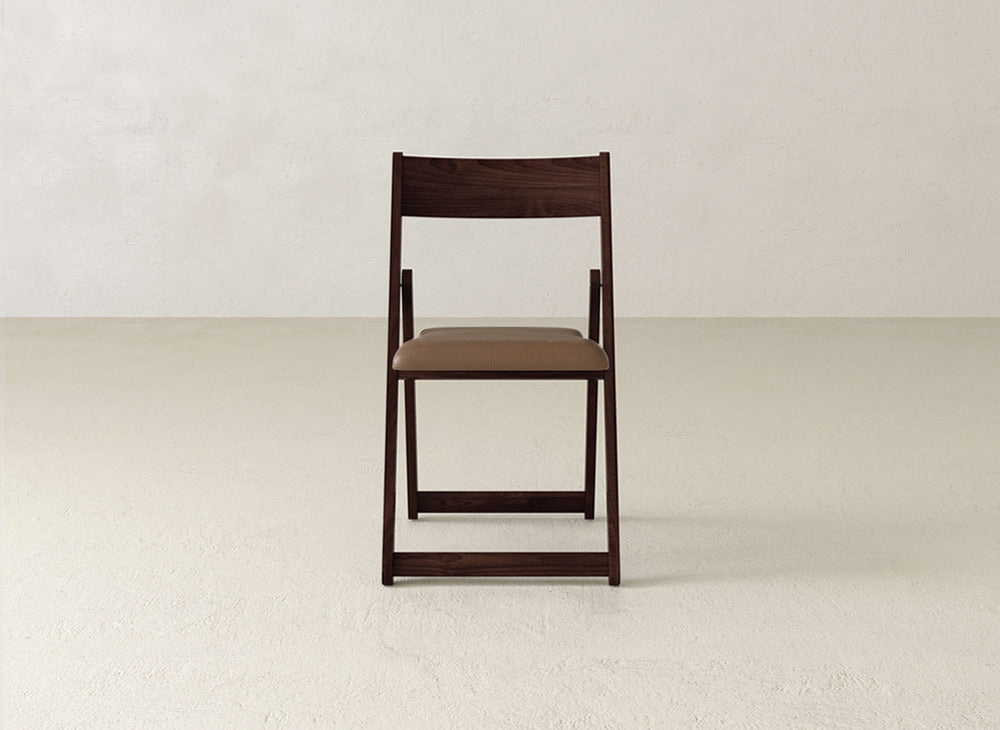 The Emmett Upholstered Folding Chair by James & James, featuring a brown padded seat and solid maple hardwood frame, sits on a light floor against an off-white wall.
