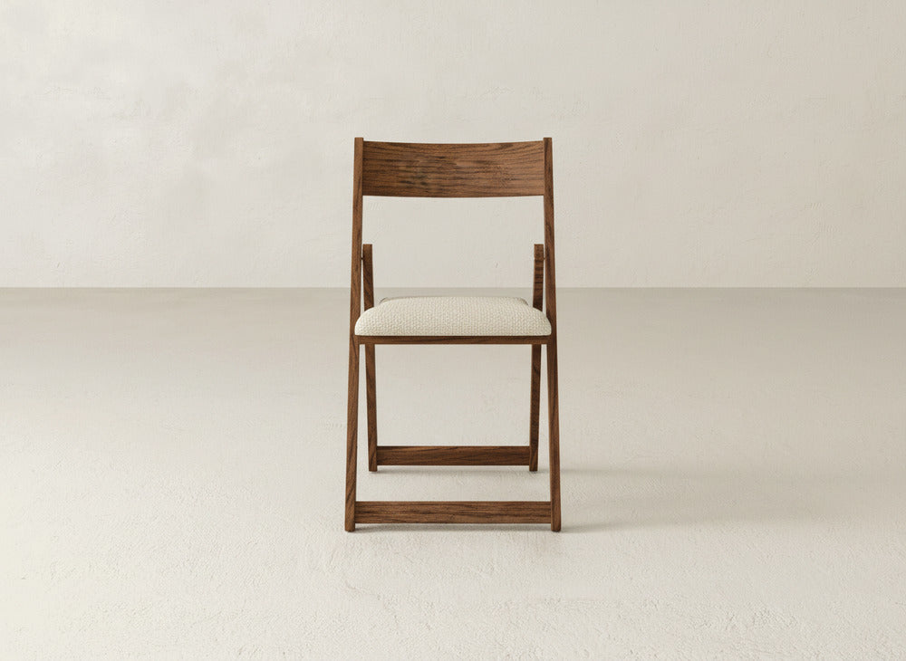 The Emmett Upholstered Folding Chair by James & James, crafted from solid maple hardwood with a light cushioned seat, stands centered in a minimalist room, showcasing its elegant and functional foldable design.