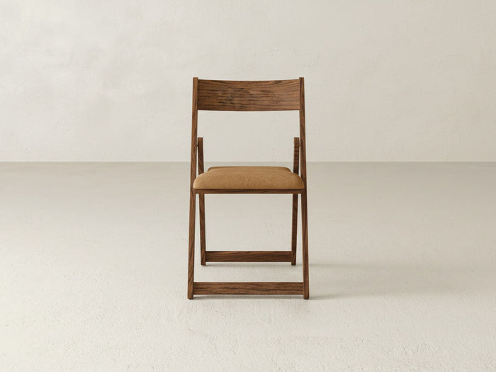 The Emmett Upholstered Folding Chair by James & James, featuring a tan cushioned seat, sits in the center of a plain, light-colored room, highlighting its versatile foldable design against a minimal backdrop.