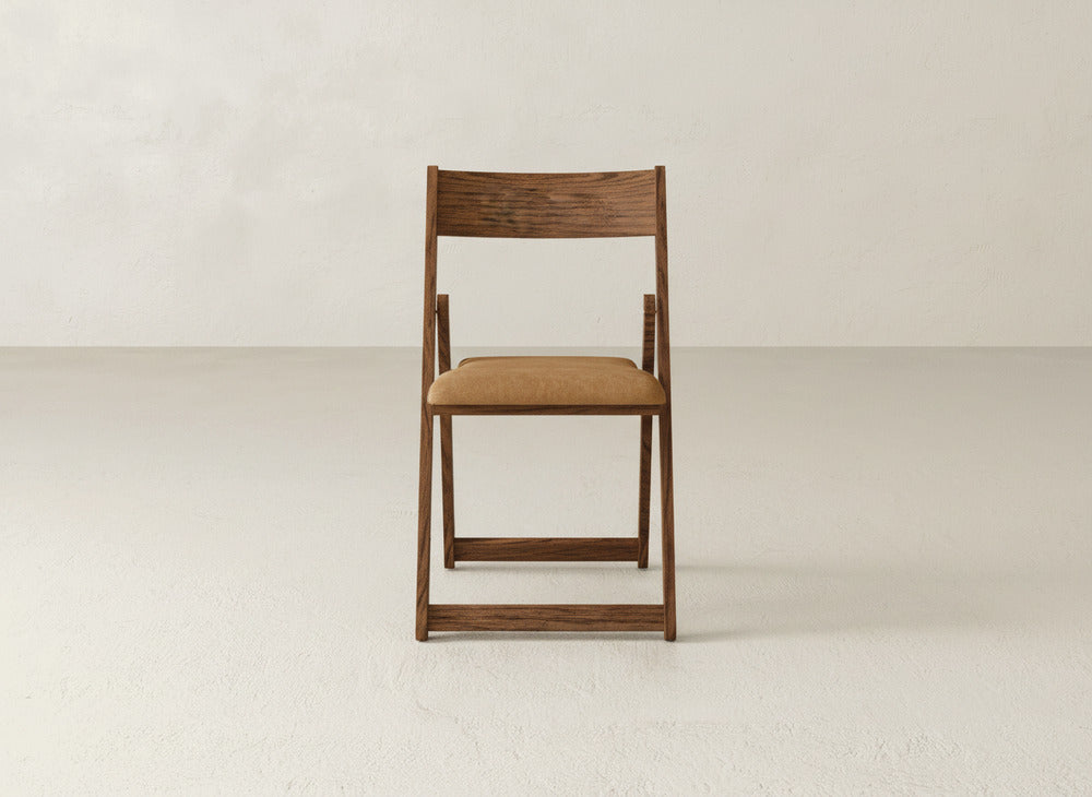 The Emmett Upholstered Folding Chair by James & James, featuring a tan cushioned seat, sits in the center of a plain, light-colored room, highlighting its versatile foldable design against a minimal backdrop.