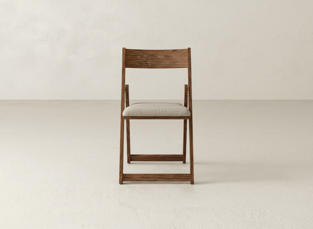The James & James Emmett Upholstered Folding Chair, made of solid maple hardwood with a light beige cushioned seat, stands upright against a plain, light-colored wall and floor.