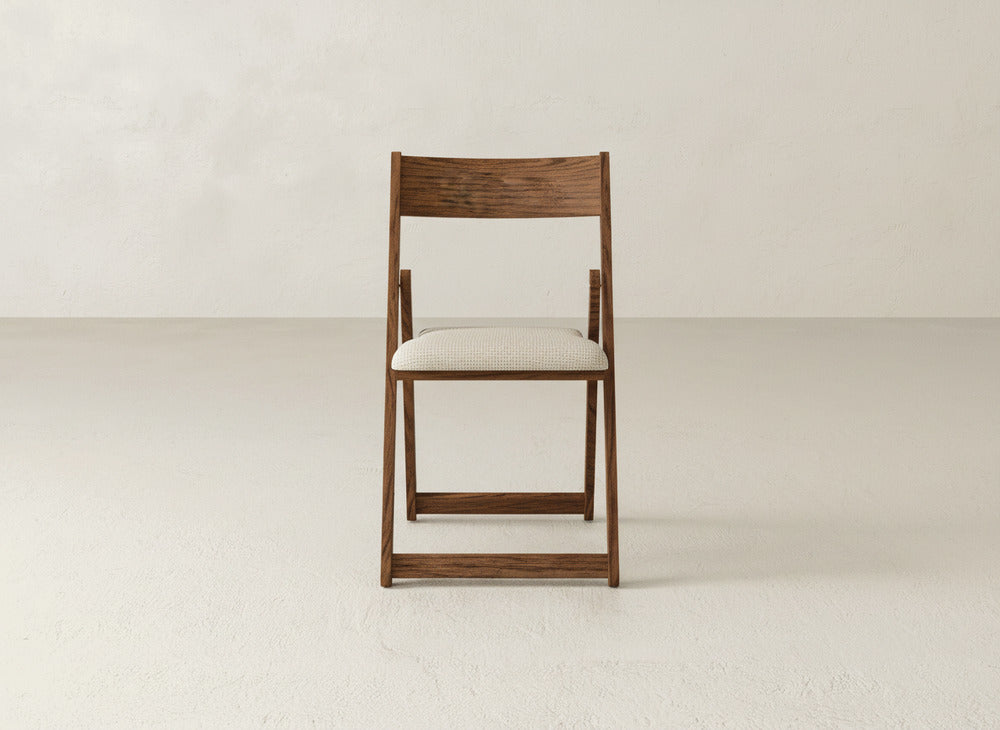 The Emmett Upholstered Folding Chair by James & James, featuring a maple hardwood frame and a light cushioned seat, is set on a plain, light floor against a minimal off-white wall.