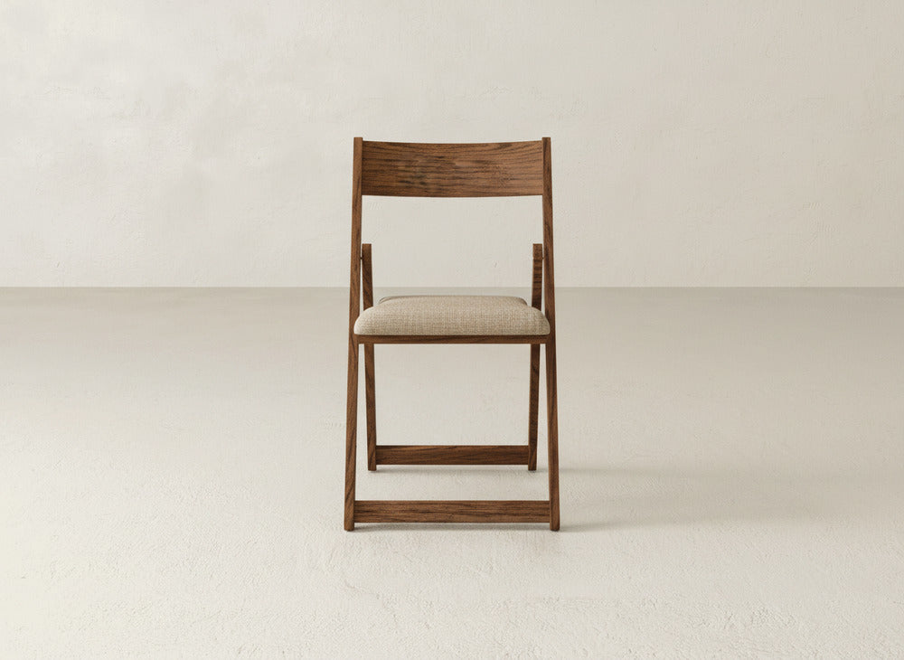 The Emmett Upholstered Folding Chair by James & James, featuring a solid maple hardwood frame and light beige cushioned seat, sits in the center of a simple, light-toned room with a smooth floor and textured wall.