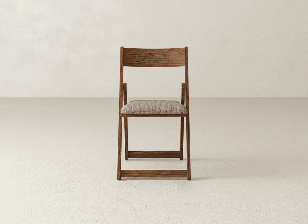 The Emmett Upholstered Folding Chair by James & James, featuring a patterned fabric cushion and solid maple frame, sits centered against a plain light wall and floor, showcasing its elegant foldable design.