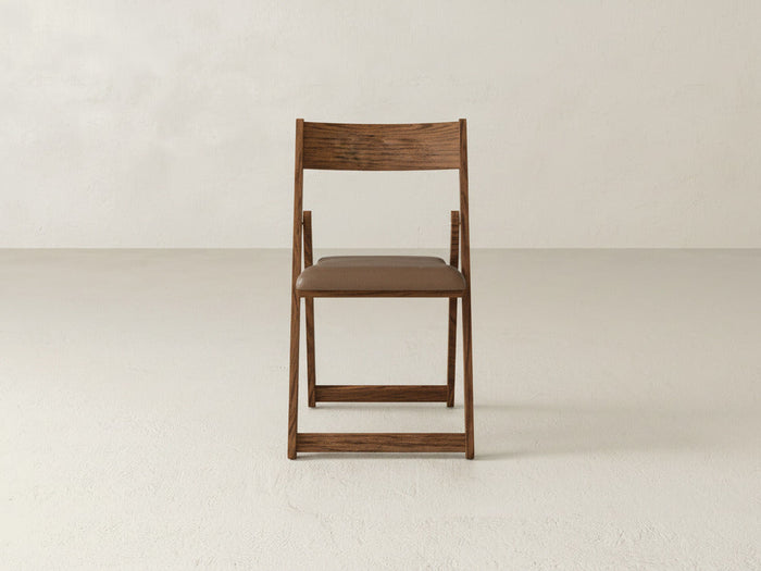 The Emmett Upholstered Folding Chair by James & James, featuring a brown seat cushion and solid maple hardwood, is centered on a light floor against an off-white wall, emphasizing its practical foldable design.