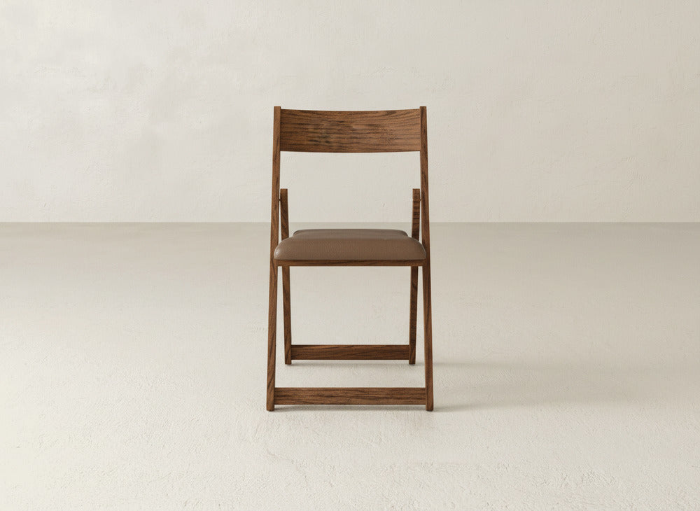 The Emmett Upholstered Folding Chair by James & James, featuring a brown seat cushion and solid maple hardwood, is centered on a light floor against an off-white wall, emphasizing its practical foldable design.