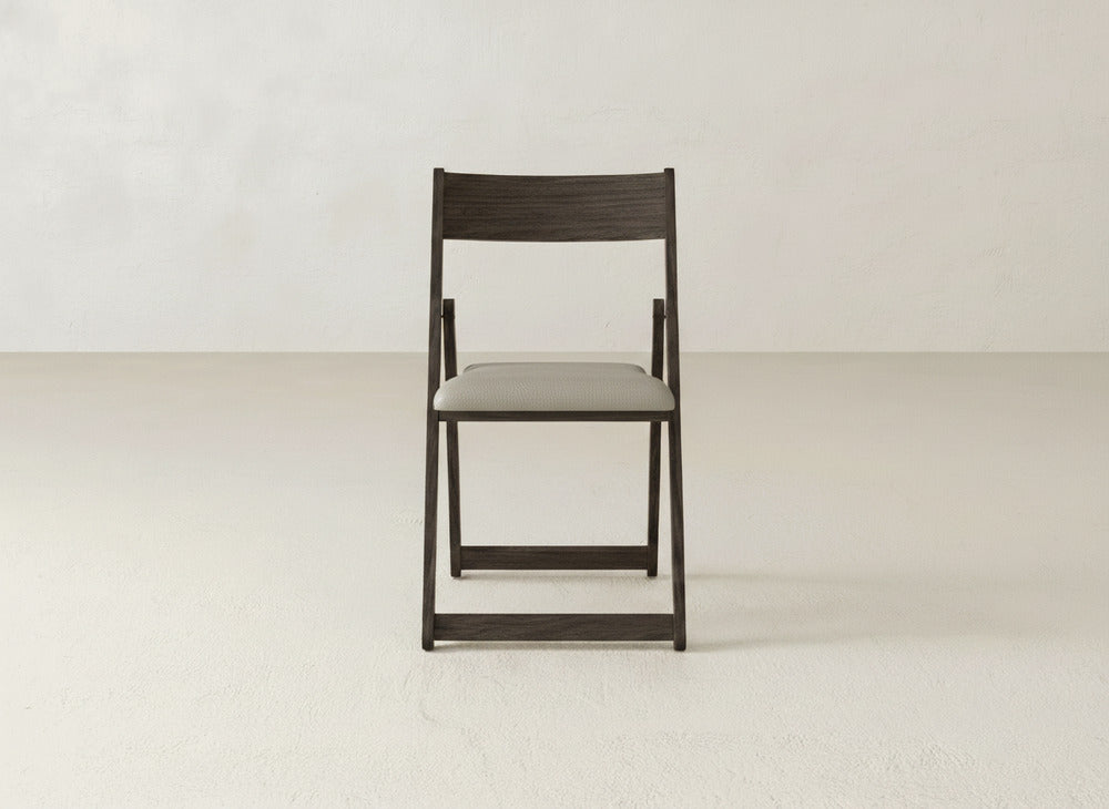 The Emmett Upholstered Folding Chair by James & James features a dark wood frame and light cushioned seat. Photographed front-on, it sits on a light floor against an off-white wall, highlighting its classic foldable design.