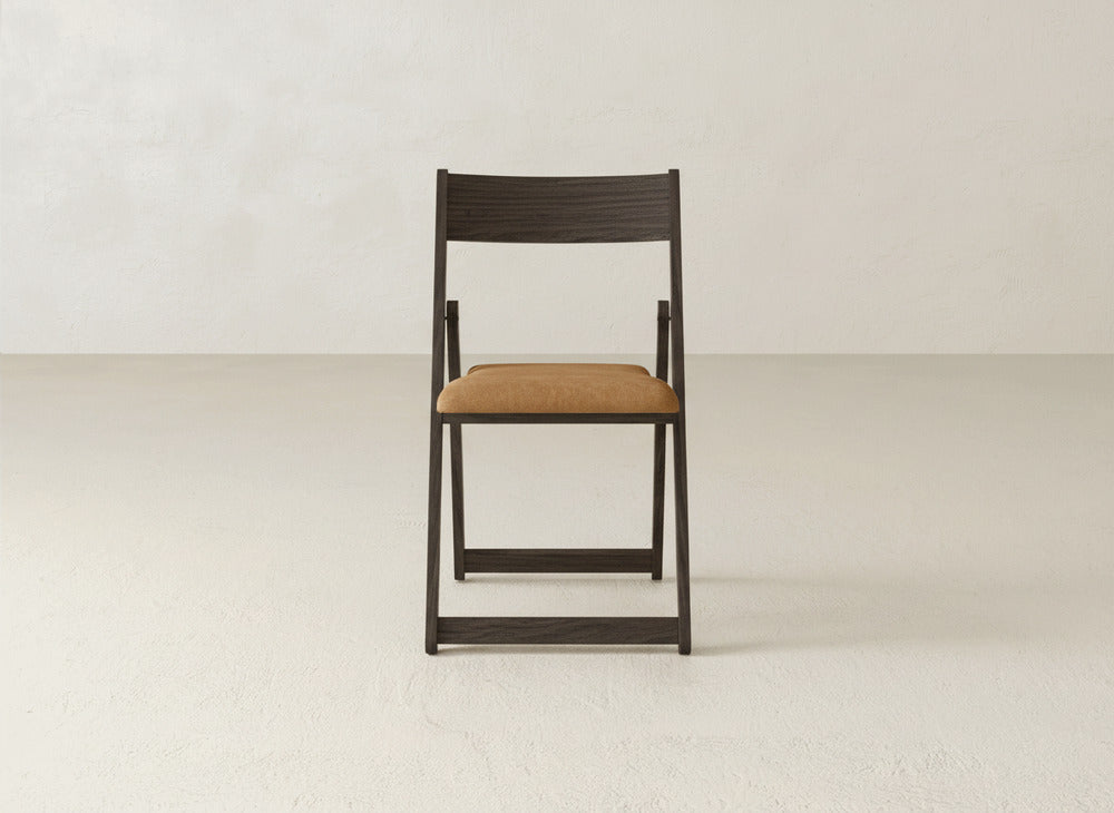 The James & James Emmett Upholstered Folding Chair, featuring dark solid maple hardwood and a tan cushioned seat, is centered against a plain, light-colored wall and floor.
