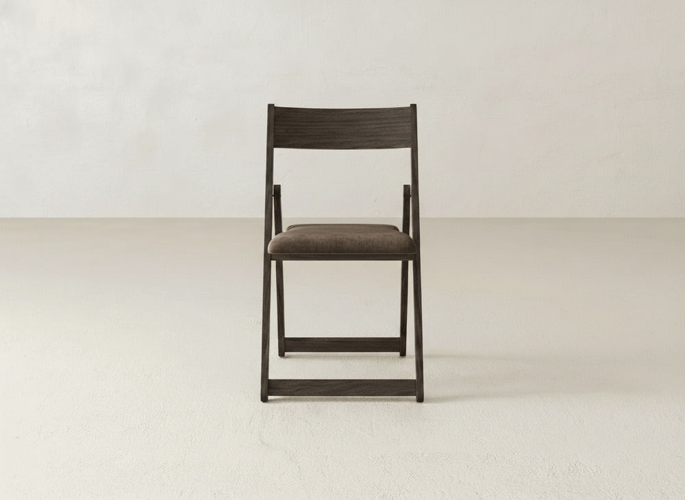 The James & James Emmett Upholstered Folding Chair, featuring a dark wood frame and cushioned brown seat, is centered in a minimalist room with light walls and a simple, light-colored floor.