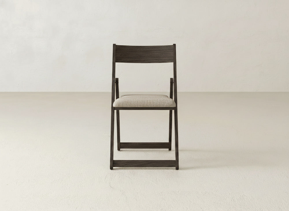 The James & James Emmett Upholstered Folding Chair, featuring a dark frame and light cushioned seat, is centered in an empty, light-toned room with a smooth floor and plain wall.