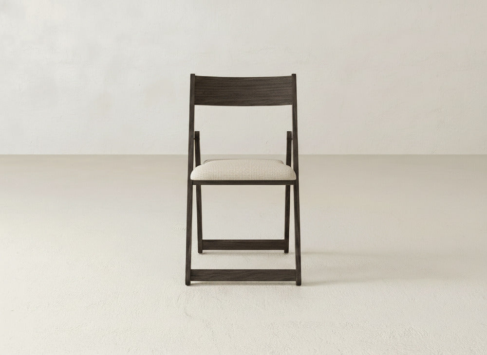 The Emmett Upholstered Folding Chair by James & James features a dark wood frame and light cushioned seat, shown facing forward in a minimalist room with a white wall and light floor to highlight its stylish, foldable design.