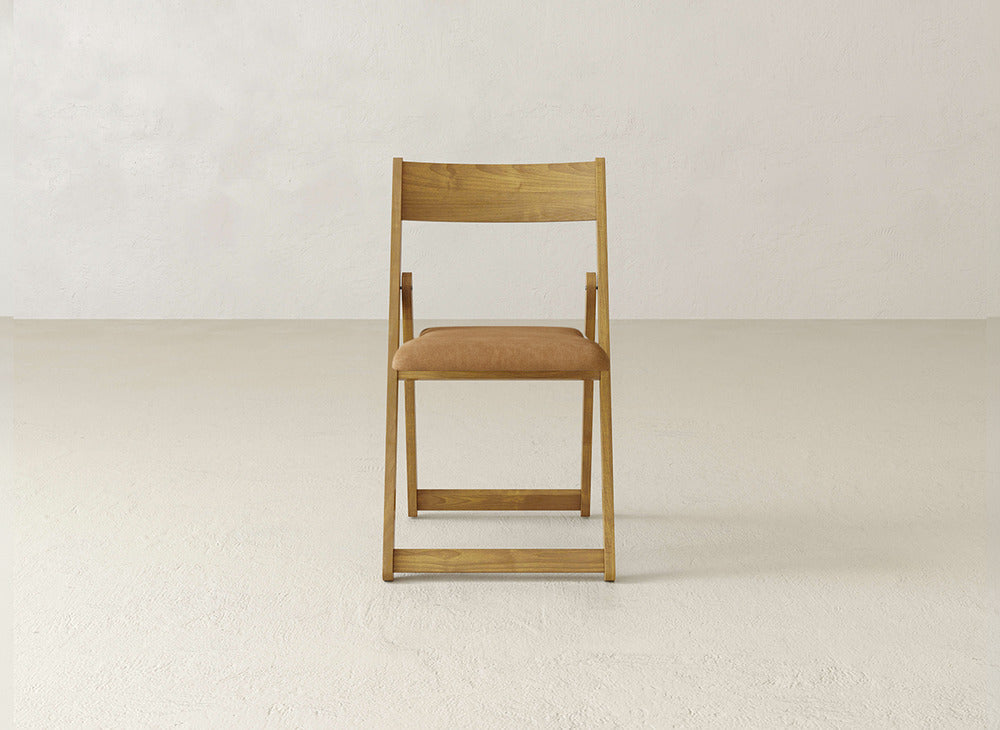 The James & James Emmett Upholstered Folding Chair, featuring a light brown cushioned seat, is positioned facing forward in an empty, light-colored room with a plain wall and floor.