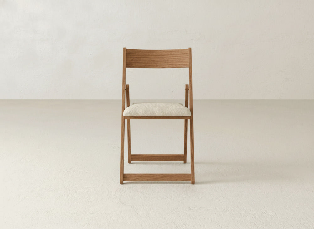 The Emmett Upholstered Folding Chair by James & James, featuring a light-colored cushioned seat and solid maple hardwood frame, is centered in a minimalist room with white walls and a light floor.