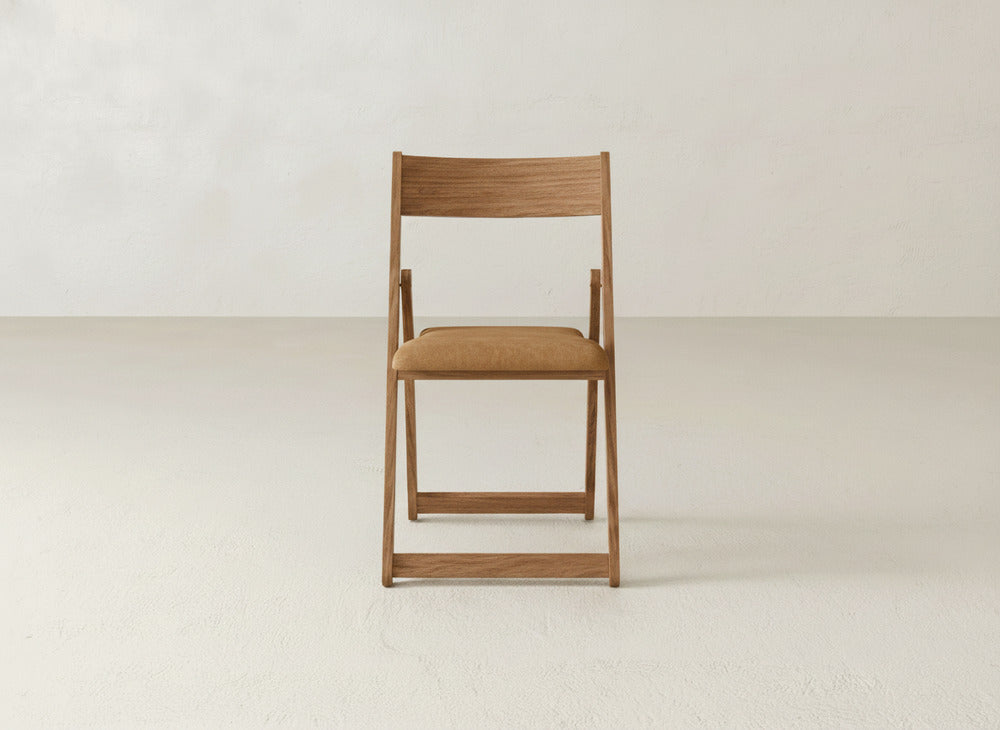 The Emmett Upholstered Folding Chair by James & James, made from solid maple hardwood with a light brown cushioned seat, is shown facing forward in an empty room with a neutral background.