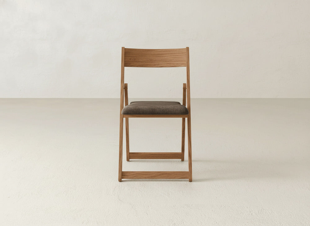 The Emmett Upholstered Folding Chair by James & James, featuring a solid maple hardwood frame and padded dark brown seat, is showcased against a light wall and floor, highlighting its elegant, foldable design.