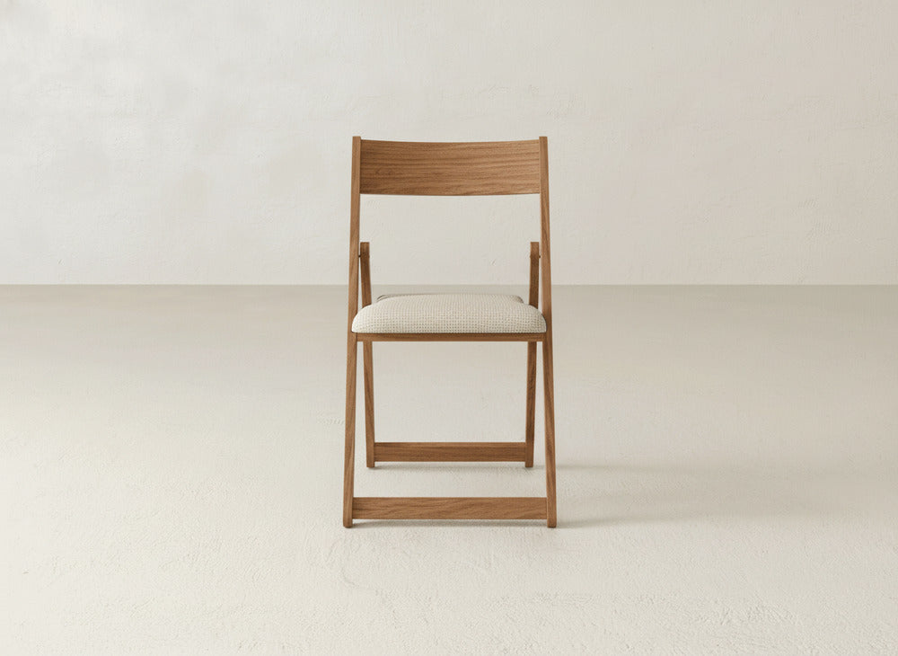 The James & James Emmett Upholstered Folding Chair, centered in a minimalist room with light floors and walls, features elegant maple hardwood and a cushioned seat that showcases its stylish, space-saving foldable design.