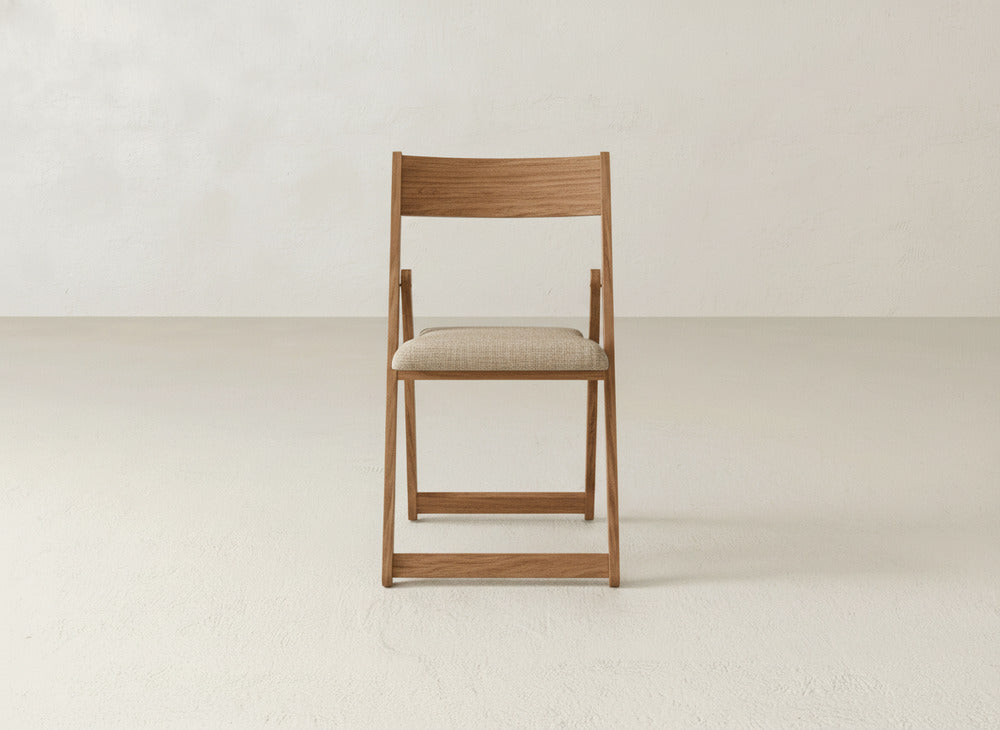 The James & James Emmett Upholstered Folding Chair, featuring a light beige cushioned seat and solid maple hardwood frame, is showcased at the center of a simple room with bare walls and a textured floor, emphasizing its elegant foldable design.