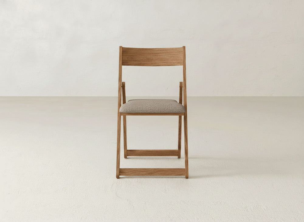 The Emmett Upholstered Folding Chair by James & James features a solid maple hardwood frame and light brown cushioned seat. Shown folded on a plain, light floor against an off-white wall, the chair faces forward.