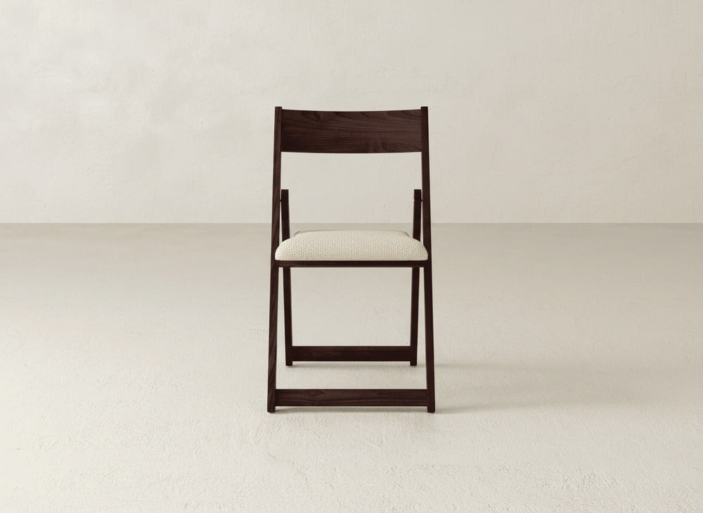 The Emmett Upholstered Folding Chair by James & James, featuring a dark wood frame and light fabric seat, is shown front-facing on a plain light floor with a textured off-white wall in the background.