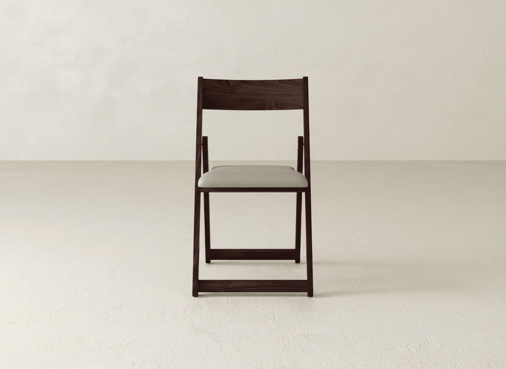 The Emmett Upholstered Folding Chair by James & James features dark wood and a light cushioned seat, shown on a plain light floor against a neutral textured wall.