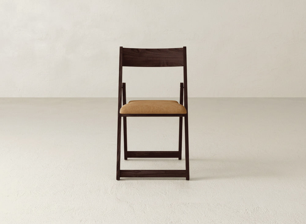 The Emmett Upholstered Folding Chair by James & James, featuring a dark brown frame and light tan cushioned seat, is positioned facing forward in an empty, light-colored room.
