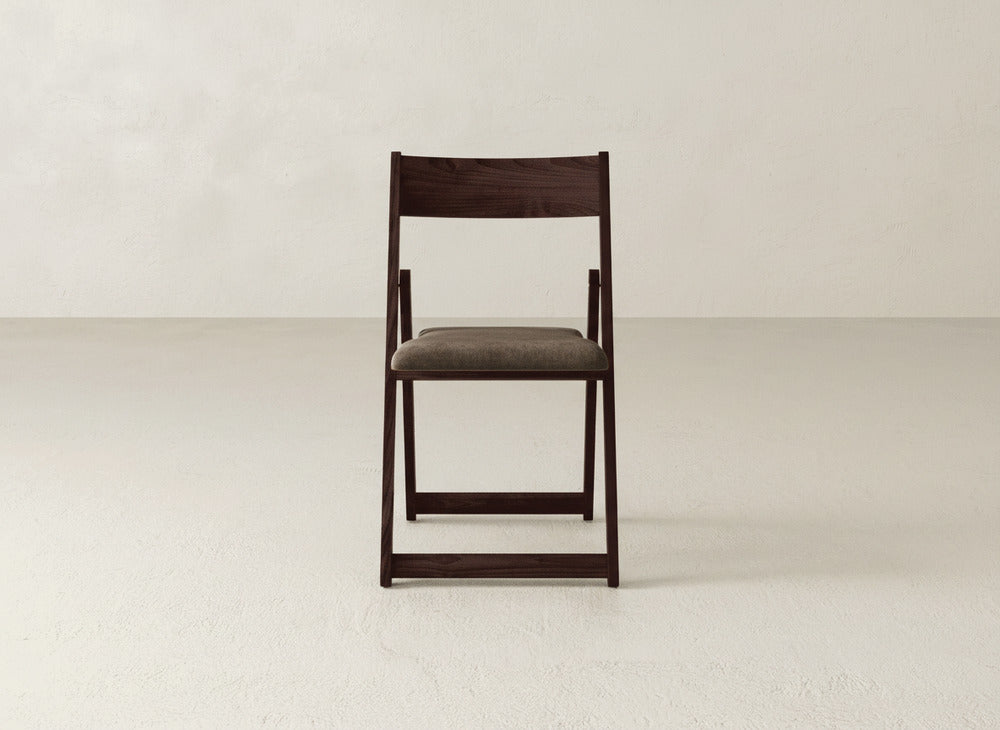 The Emmett Upholstered Folding Chair by James & James, featuring dark maple hardwood and a padded gray seat, stands in the center of a light, empty room with a subtly textured wall.