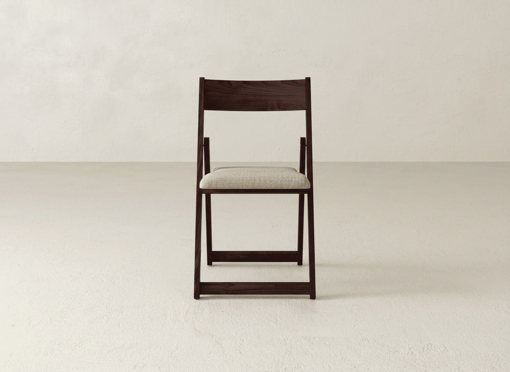 The Emmett Upholstered Folding Chair by James & James, featuring dark wood and a light beige cushioned seat, sits on a plain light floor against a simple off-white wall.