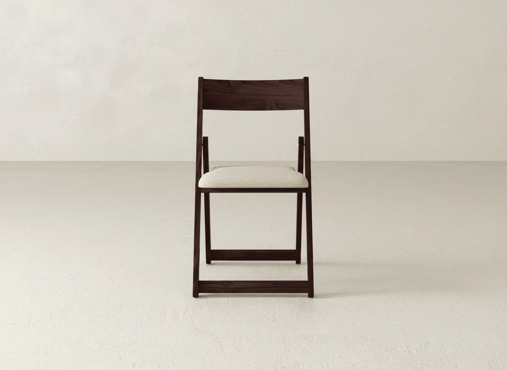 The Emmett Upholstered Folding Chair by James & James features a simple dark maple hardwood frame and a light cushioned seat, shown facing forward against a plain, light-colored wall and floor.