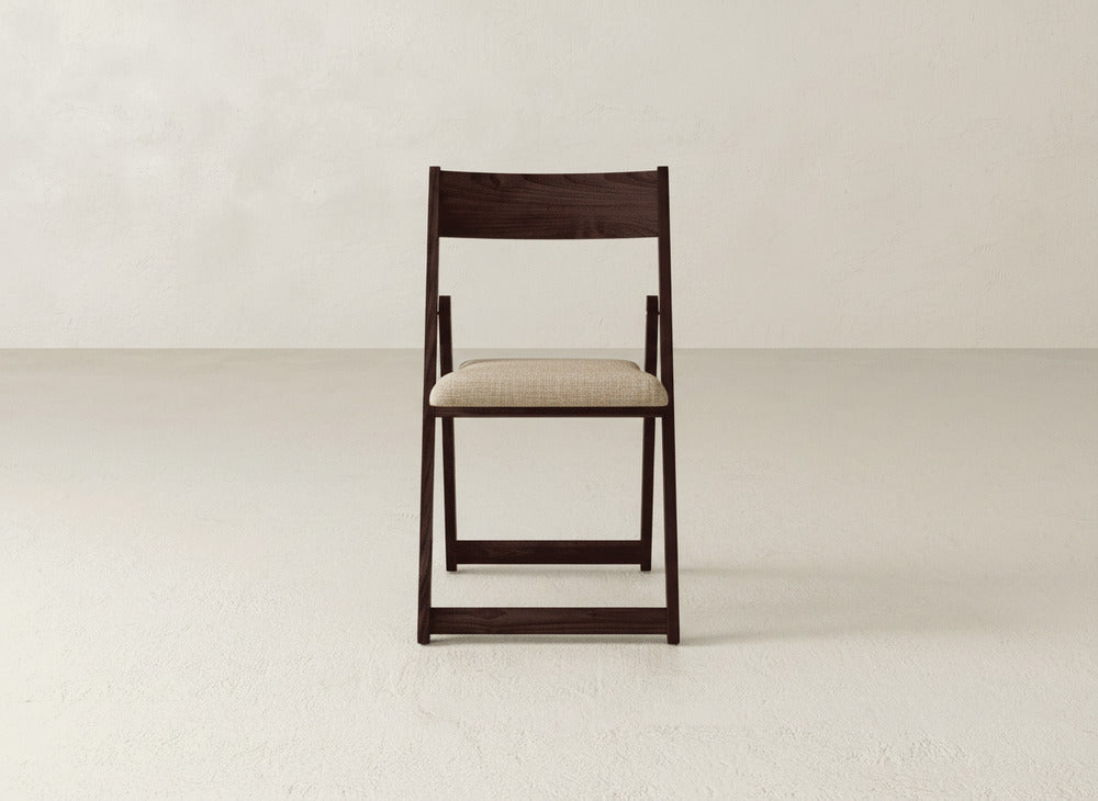 The Emmett Upholstered Folding Chair by James & James features a dark wooden frame and a light beige cushioned seat, shown facing forward against a plain, light backdrop.