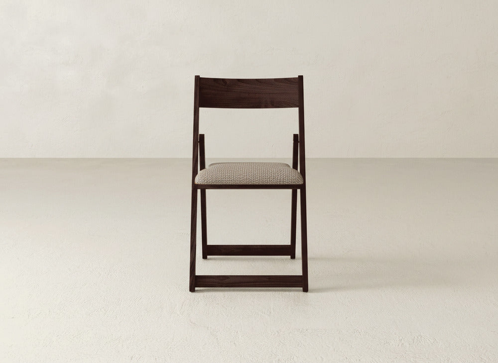 The Emmett Upholstered Folding Chair from James & James, featuring a maple hardwood frame and light textured seat cushion, is centered in an empty room with a neutral, textured wall and floor.