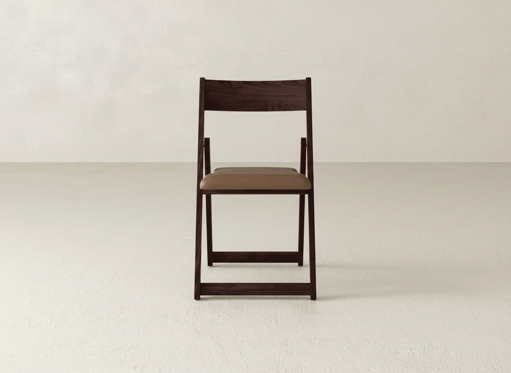 The Emmett Upholstered Folding Chair by James & James, featuring dark maple hardwood and a brown padded seat, is centered and facing forward on a light textured floor against a plain off-white wall.
