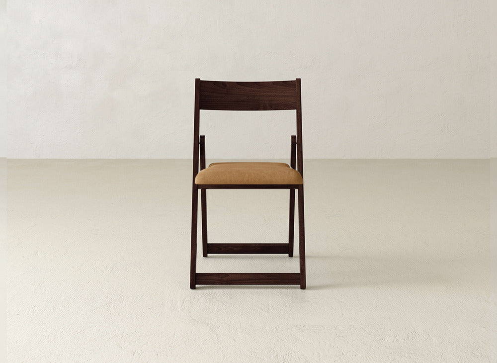 The Emmett Upholstered Folding Chair by James & James, featuring a brown cushioned seat and solid maple hardwood frame, sits against a plain textured white wall atop a light-colored floor.