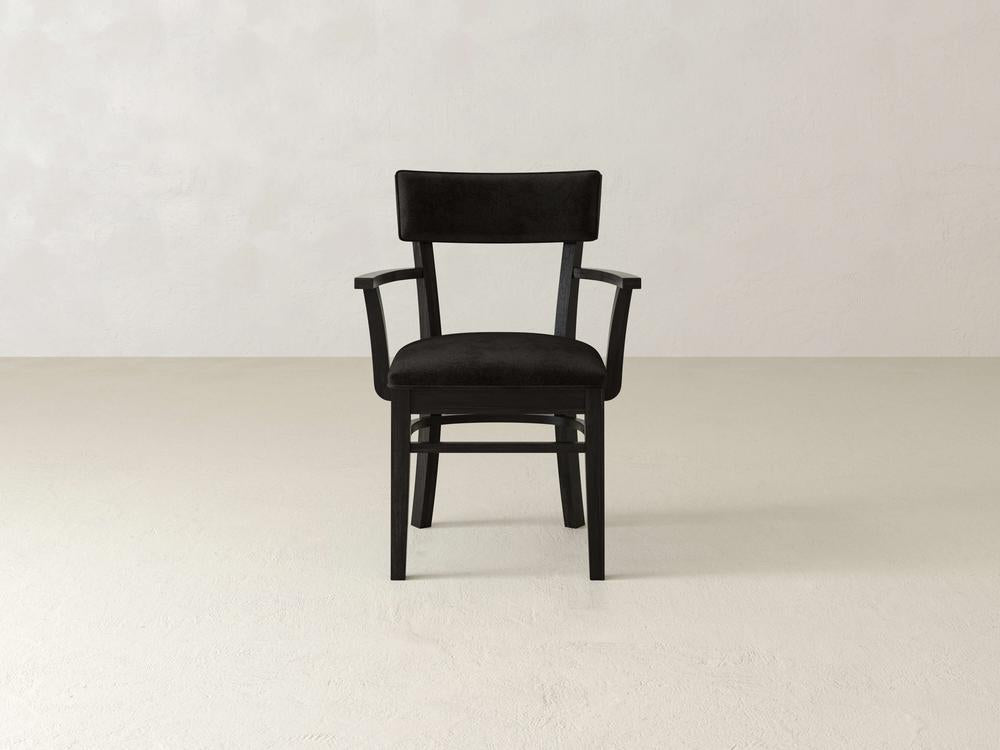 A black wooden chair with a cushioned seat and backrest, featuring armrests, is placed on a beige floor against a light gray wall. The chair is centered in the frame, highlighting its simple and elegant design.