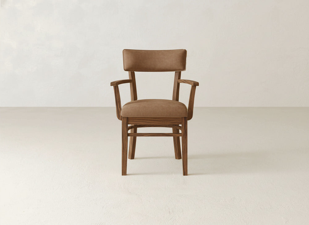 The Everly Arm Dining Chair by James+James features a cushioned brown seat, backrest, and armrests, elegantly centered against a plain, light-colored background.
