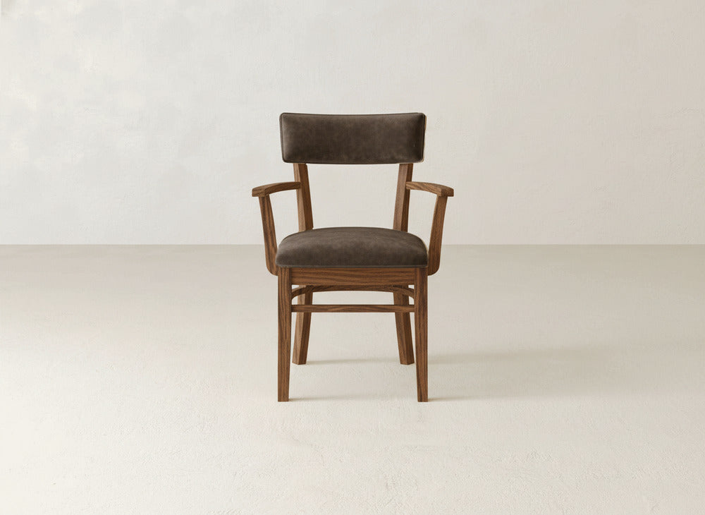 The Everly Arm Dining Chair by James+James features a dark brown cushioned seat and curved armrests, shown on a light-colored floor against an off-white wall.