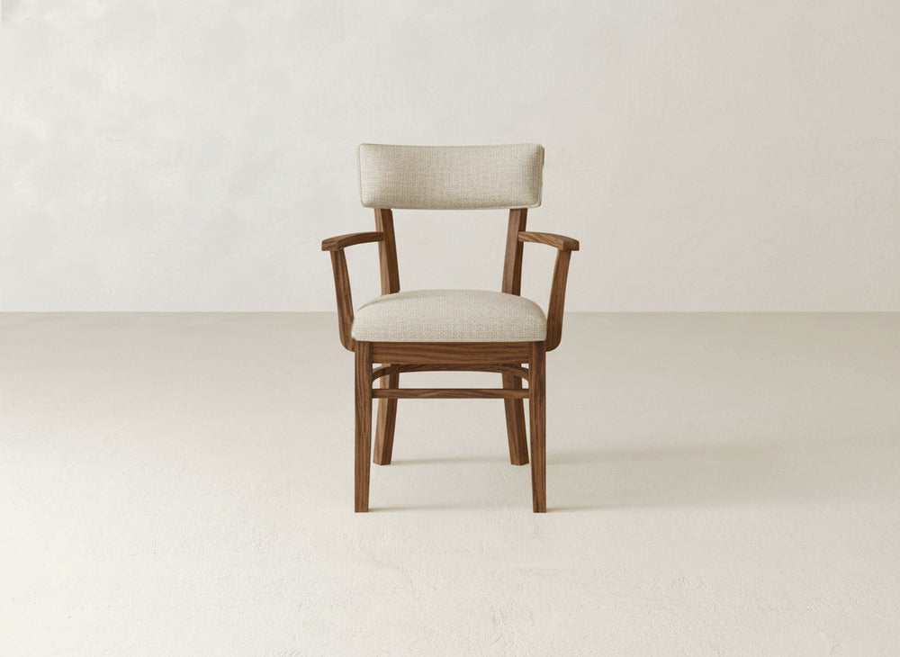 The Everly Arm Dining Chair by James+James features wooden armrests and a light beige cushioned seat and backrest, shown on a plain, light-colored floor against a neutral wall.