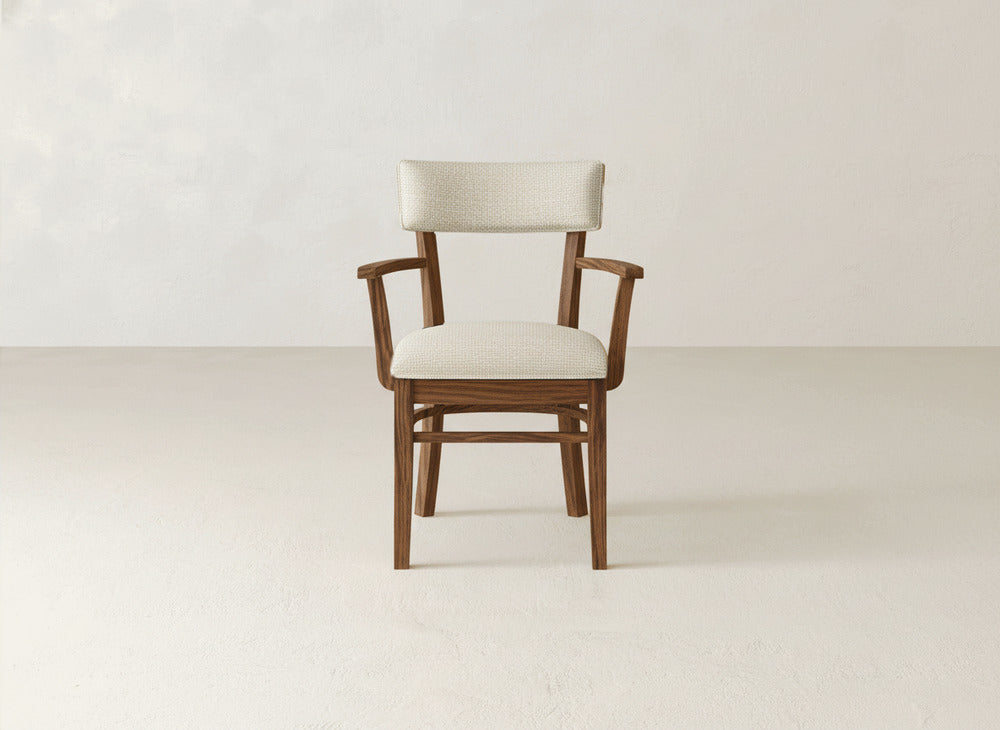 The Everly Arm Dining Chair by James+James features armrests and a light beige upholstered seat and backrest, displayed on a light-colored floor against an off-white wall for a stylish, mid-century modern look.