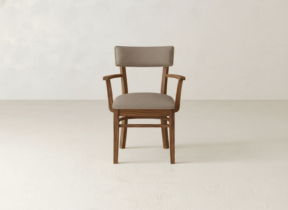 The Everly Arm Dining Chair by James+James, featuring armrests and light brown fabric upholstery with cushioned seat and back, is centered against a plain, light-colored wall and floor.