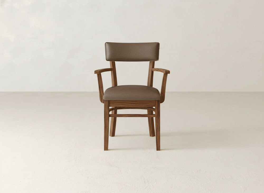 The Everly Arm Dining Chair by James+James features a brown upholstered seat and backrest, wooden frame, and armrests. It stands on a light floor with a plain off-white wall as the backdrop.