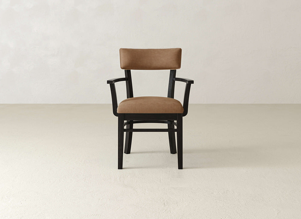 The Everly Arm Dining Chair by James+James features a brown cushioned seat and backrest, black frame, and armrests. It sits on a light smooth floor against an off-white wall, showcasing its mid-century modern style.
