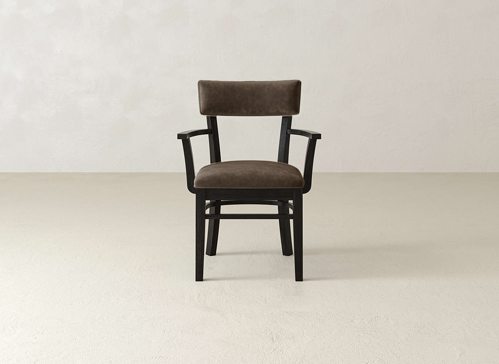 The Everly Arm Dining Chair by James+James features dark wood and a padded brown leather seat and backrest, set on a light textured floor against a plain beige wall.