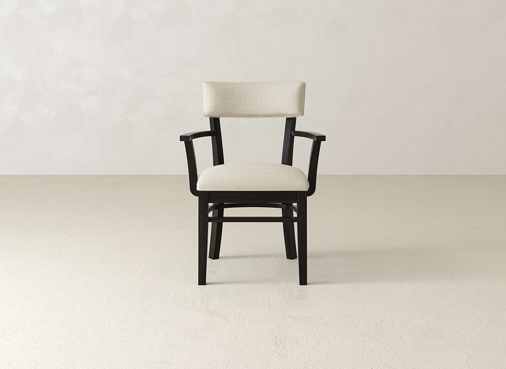 The Everly Arm Dining Chair by James+James, featuring a black frame and cream upholstered seat and backrest with armrests, is centered in a minimal, light-toned room with a textured wall and bare floor.