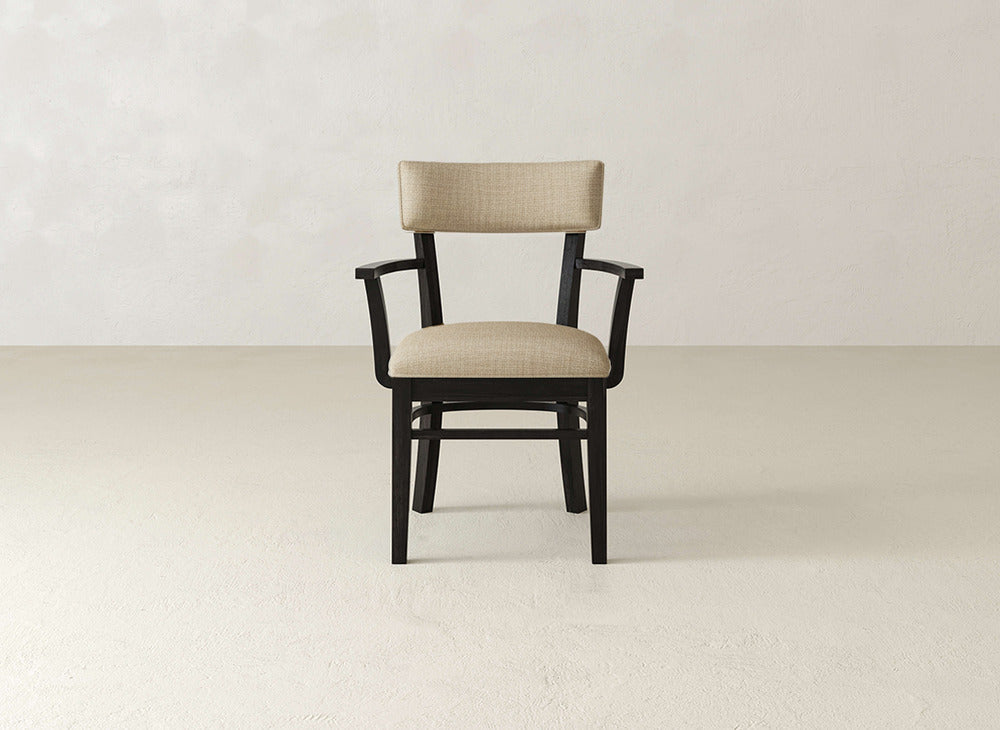 The Everly Arm Dining Chair by James+James features a black wooden frame with a beige upholstered seat and backrest, positioned on a light floor against an off-white wall.