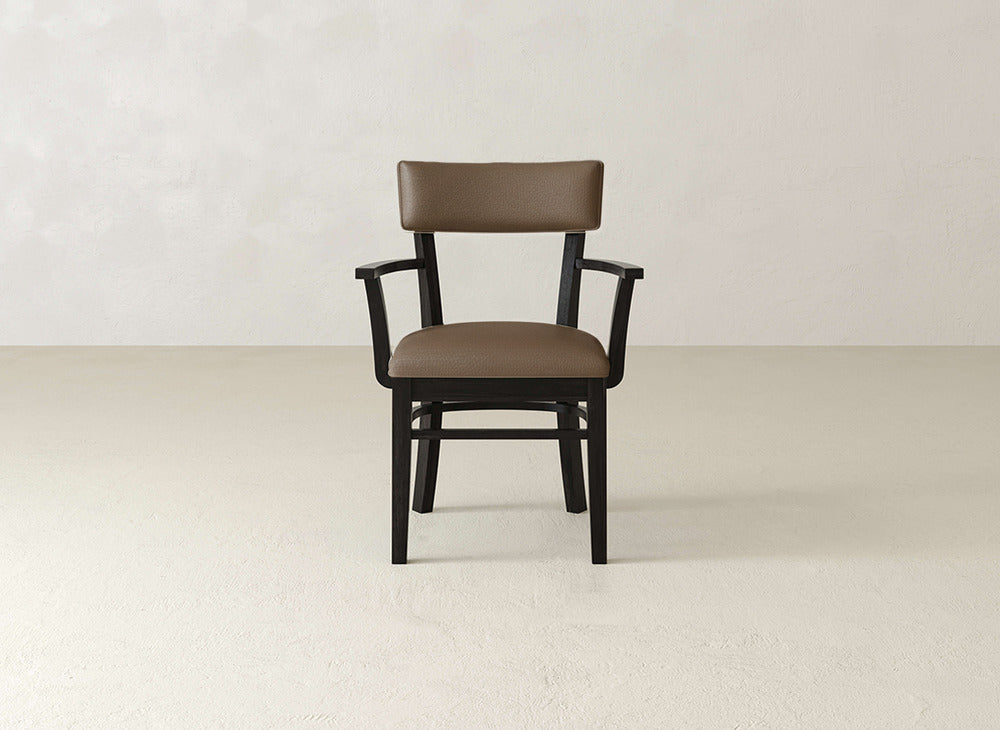 The Everly Arm Dining Chair by James+James, featuring a brown cushion, mid-century modern style, armrests, and a black wooden frame, is shown facing forward against a plain light wall and floor.