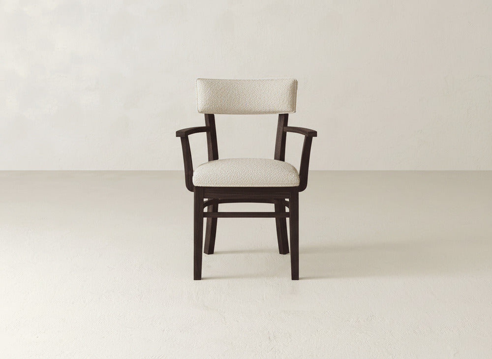 The Everly Arm Dining Chair by James+James features a modern wooden design with a dark frame and light upholstered seat and backrest, shown facing forward against a simple, light-colored wall and floor.